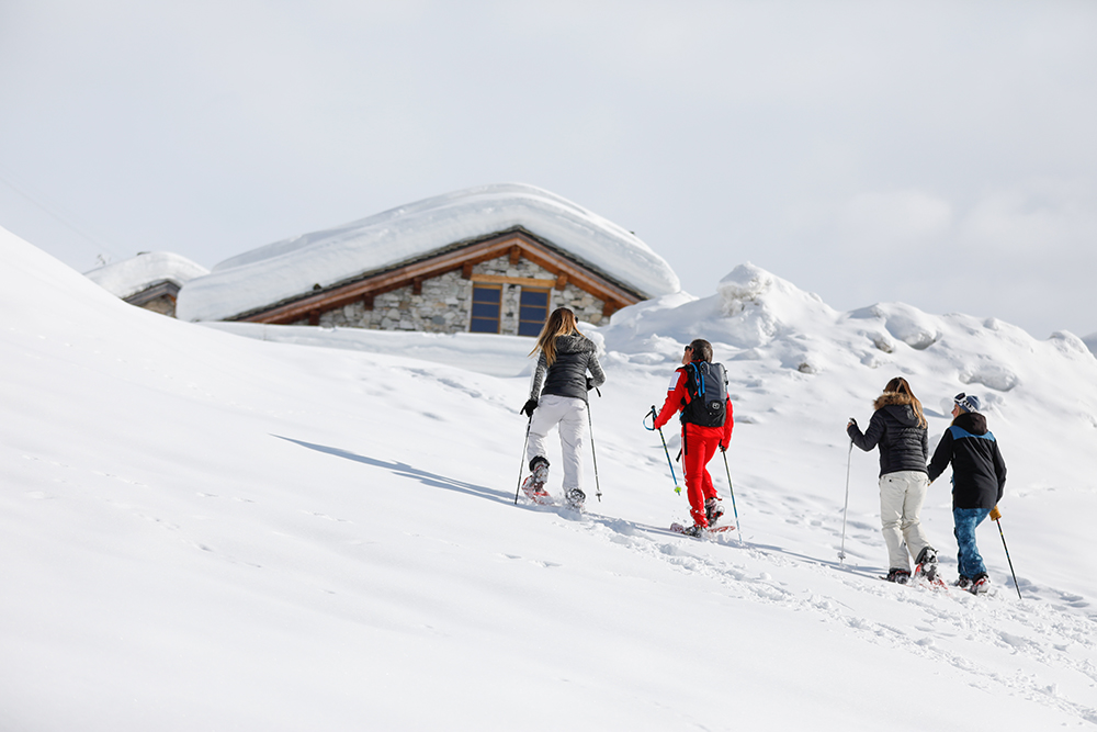 Quatre randonneurs en raquettes progressent dans la neige près d’un chalet